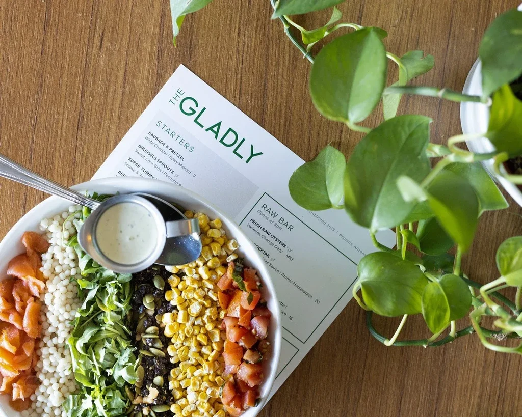 Colorful food bowl with diverse ingredients, dressing, menu on wood table.