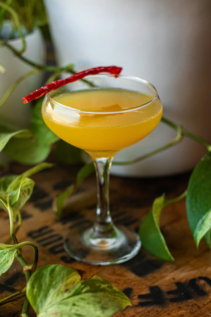Vibrant yellow cocktail with red pepper garnish, surrounded by green leaves.