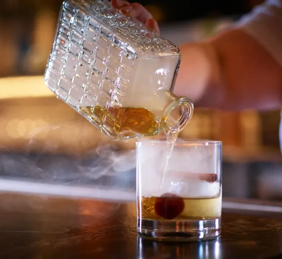Hand pours smoky amber liquid into glass with cherry, cinnamon stick.