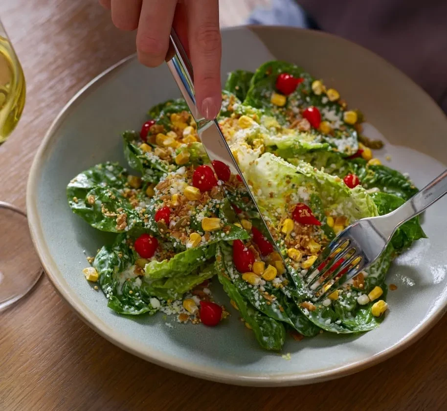 Hand cutting vibrant salad: lettuce, creamy dressing, cheese, corn, and berries.
