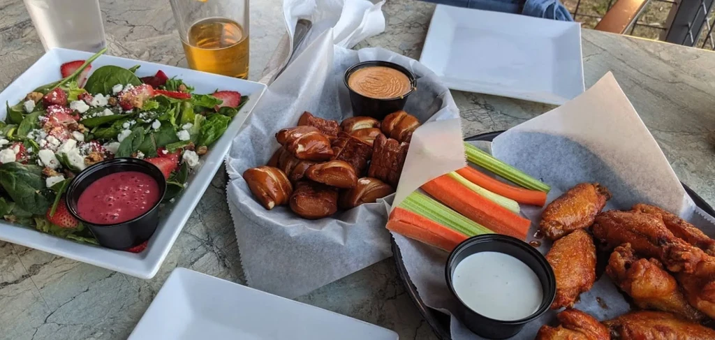 Overhead casual table with salad, pretzel bites, chicken wings, and drinks.