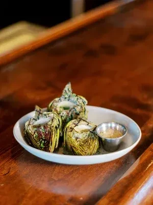 Three grilled artichoke halves with dipping sauce on a wooden table.