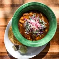 Colorful soup bowl with tortillas, lime, and spices on sunlit wood.