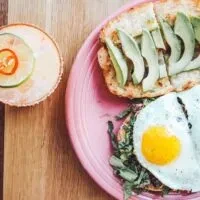 Top-down view of avocado toast, fried egg on greens, garnished drink.