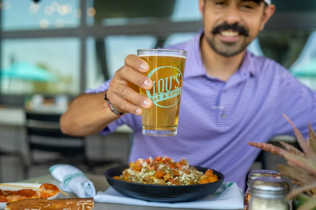 Person holds 'Lou's Bar' beer glass above a colorful food bowl.