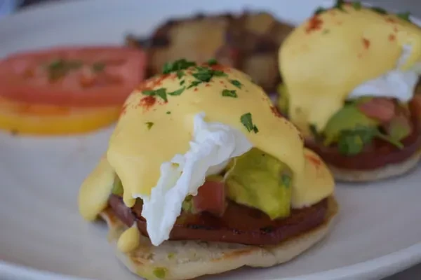 Two Eggs Benedict with runny yolk, hollandaise, avocado, and vegetables.