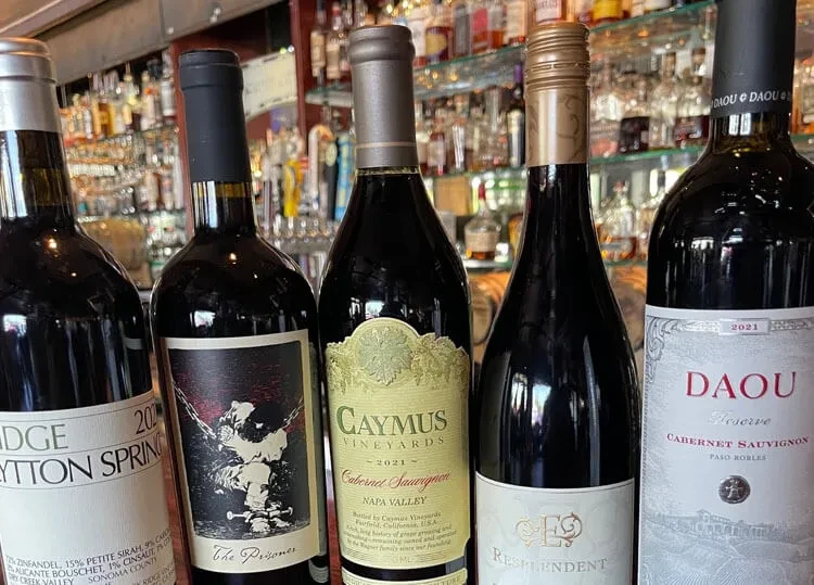 Five labeled red wine bottles lined up on a bar.