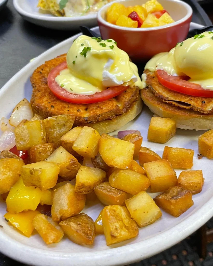 Two Eggs Benedict, roasted potatoes, and fruit salad on a plate.