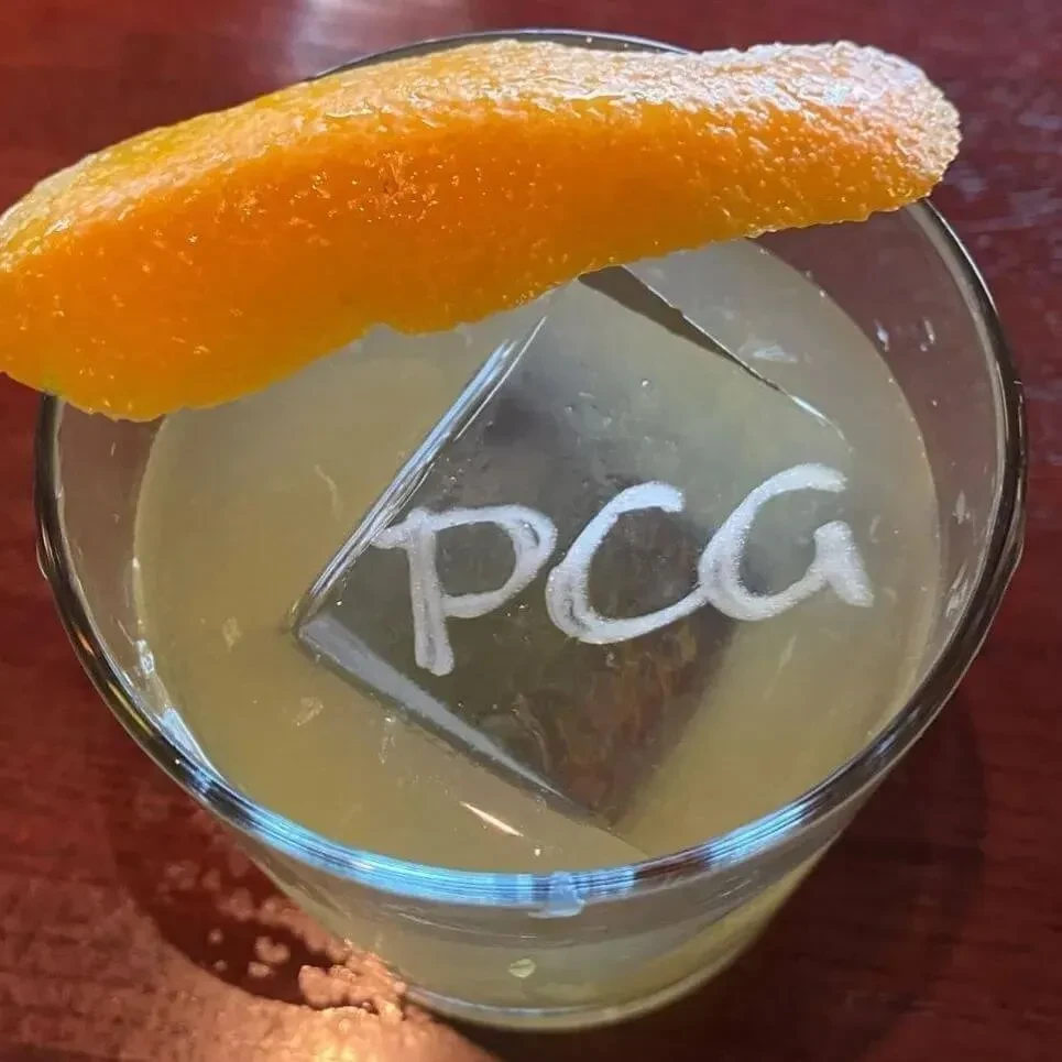 Cocktail with 'PCG' embossed ice cube and orange peel garnish.