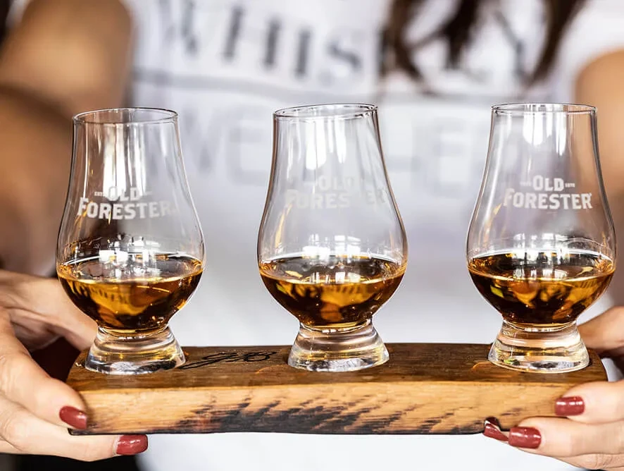 Three Old Forester whiskey glasses with amber liquid on a wooden tray.