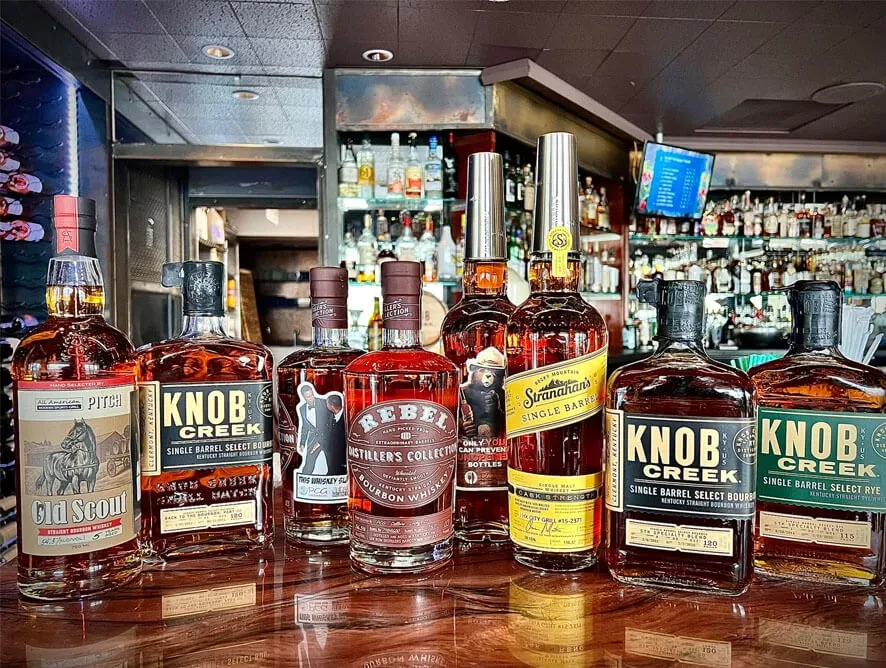 Nine whiskey and bourbon bottles on a dark bar top.