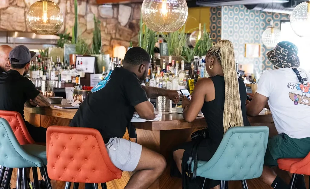 Modern bar scene with diverse patrons, vibrant stools, and warm lighting.