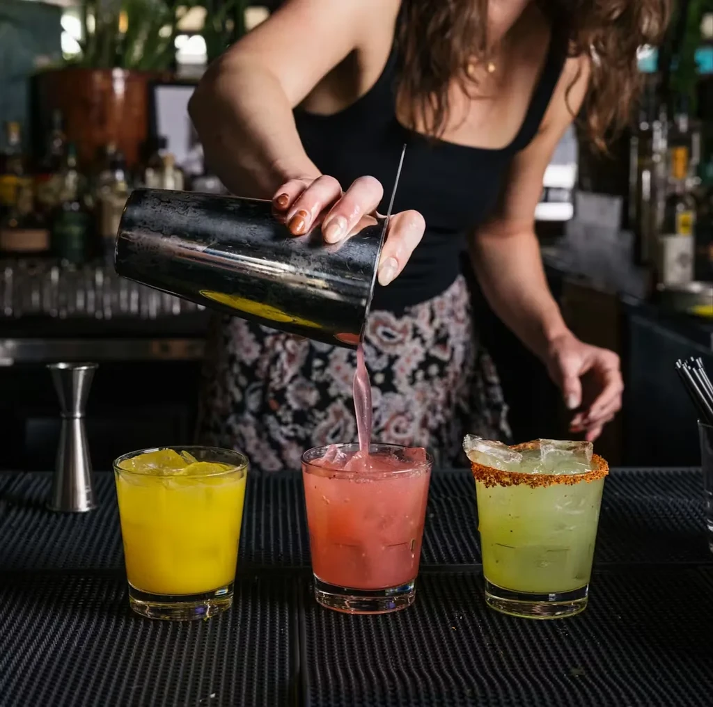 Bartender pours pink cocktail from shaker, other drinks on bar.