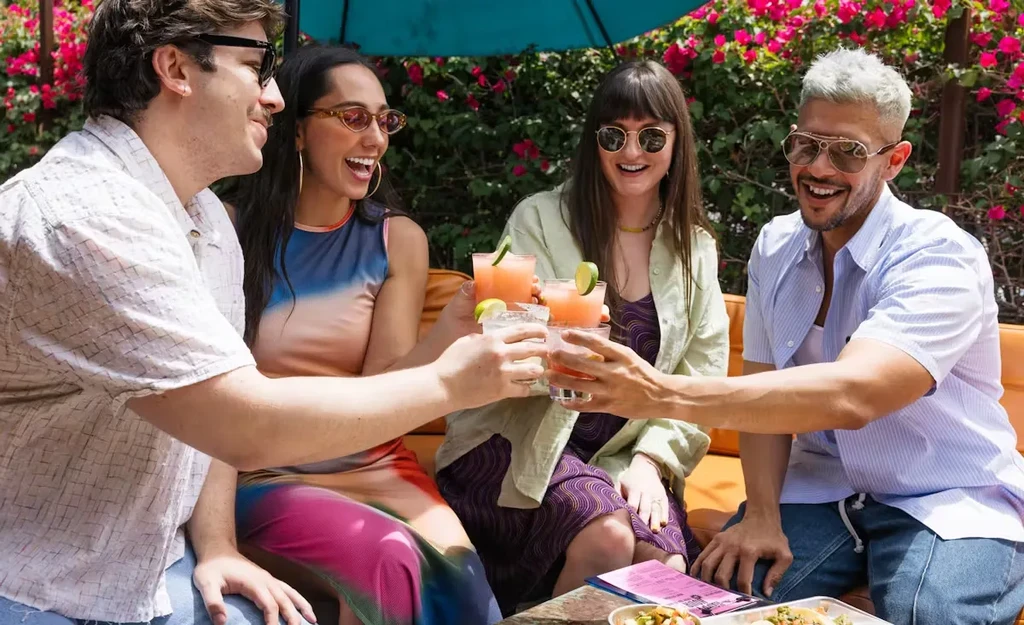Four young adults toast drinks outdoors amidst colorful flowers.