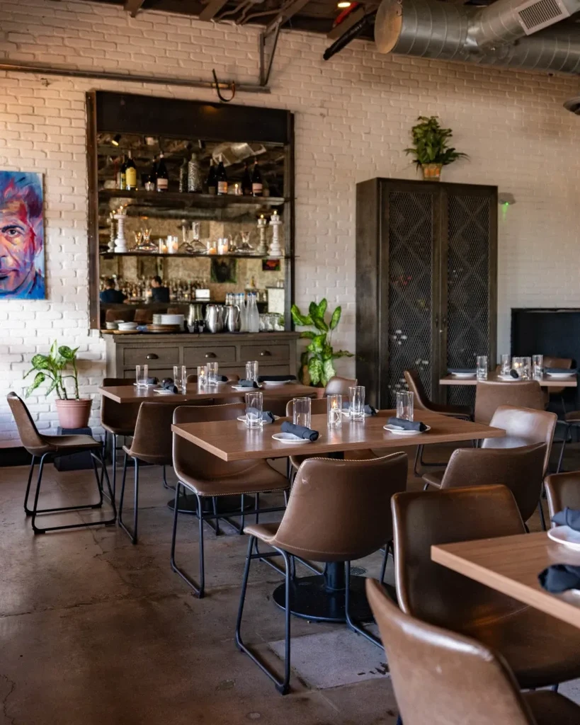 Restaurant interior with exposed brick walls, wooden tables, leather chairs, and bar.