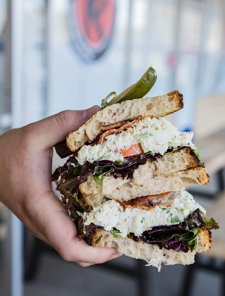 Hand holding a large sandwich with creamy salad, bacon, and pickled pepper.