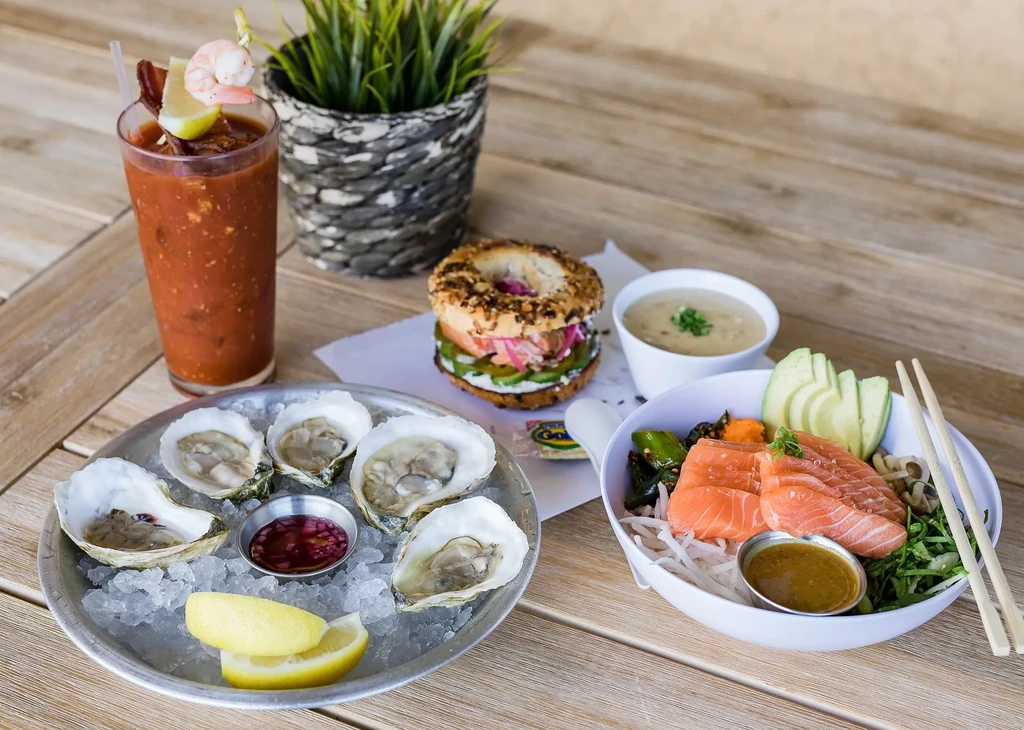 Oysters, Bloody Mary, bagel sandwich, and poke bowl on a wooden table.