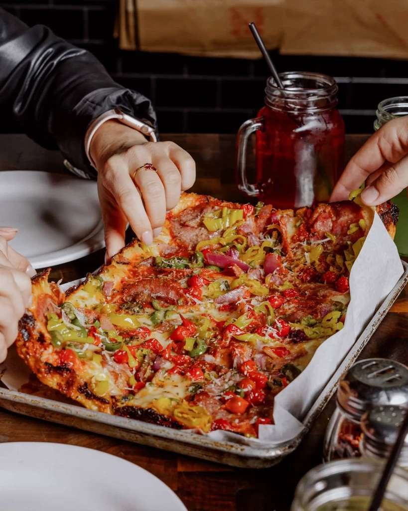 Hands sharing a rectangular pizza with colorful toppings on a tray.