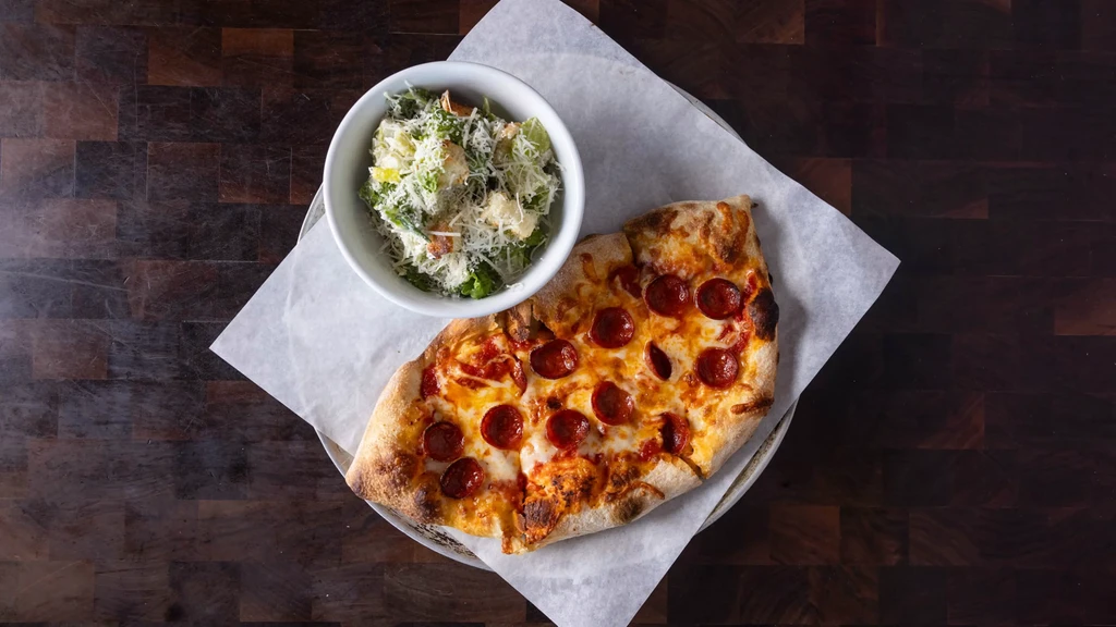 Pepperoni pizza slice and Caesar salad on a dark wooden table.