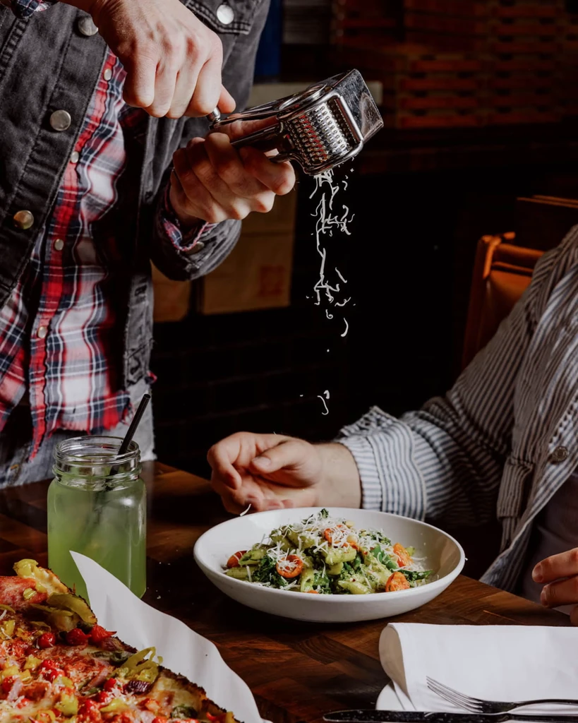 Hands grate cheese onto green pasta next to pizza and a drink.