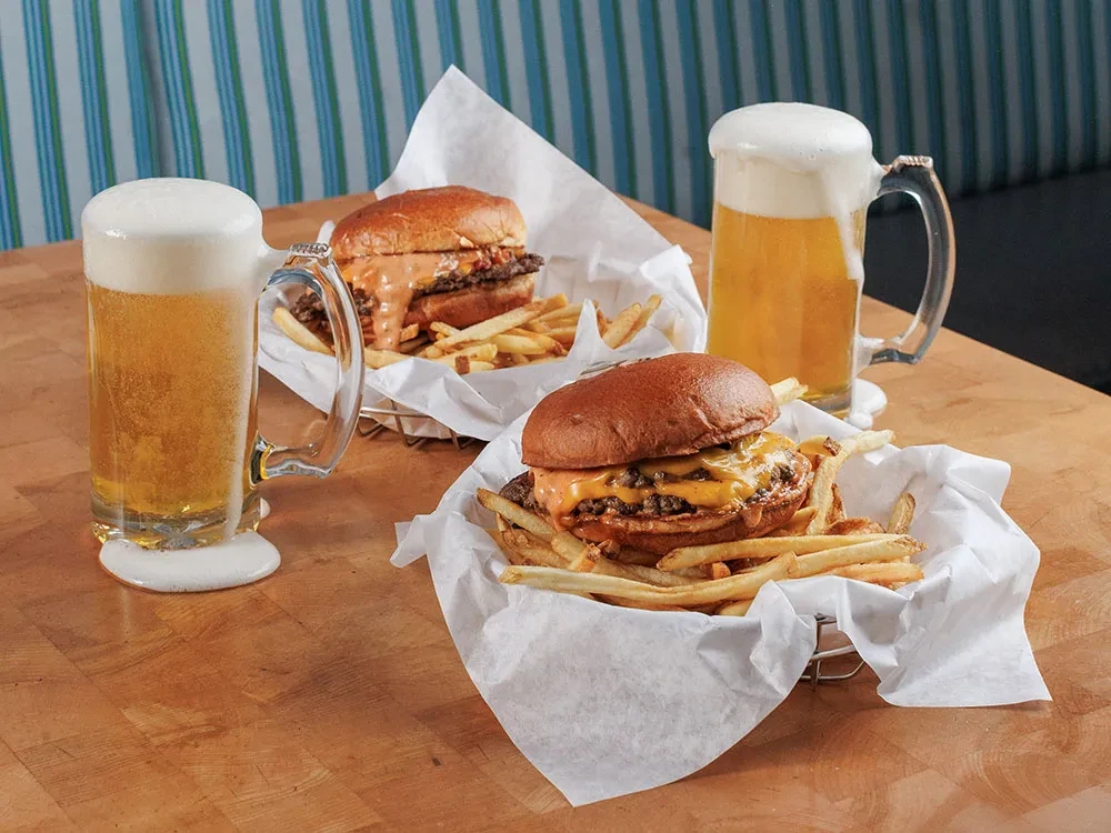 Two cheeseburgers, fries, and two beers on a wooden table.