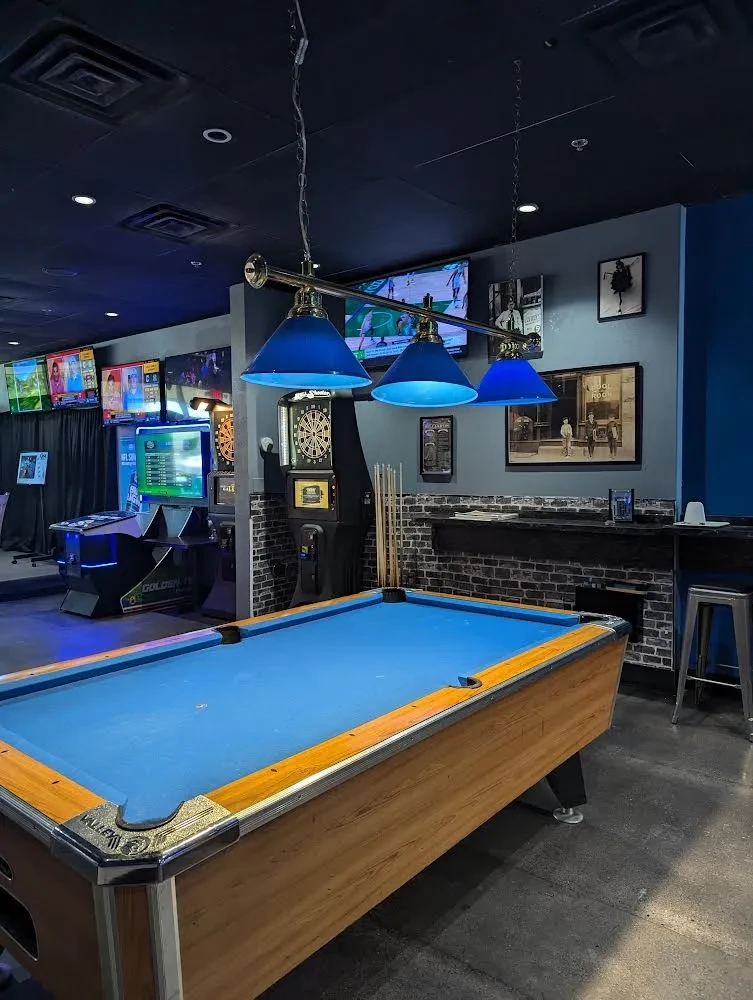 Dark game room with a blue pool table, TVs, and arcade machines.