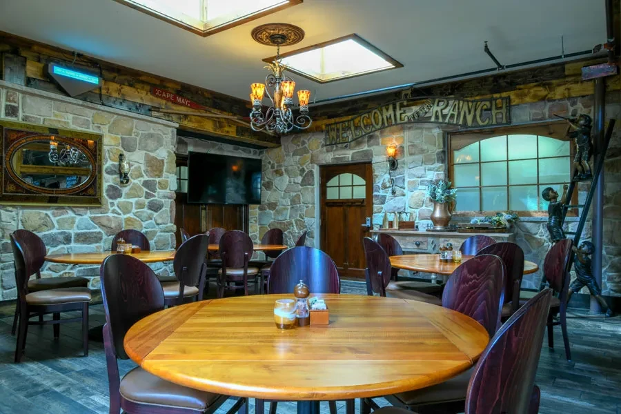 Stone-walled rustic dining room with wooden tables and "WELCOME RANCH" sign.