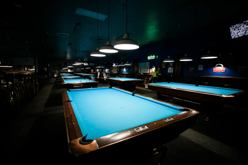 Dimly lit pool hall with turquoise tables and players under pendant lights.