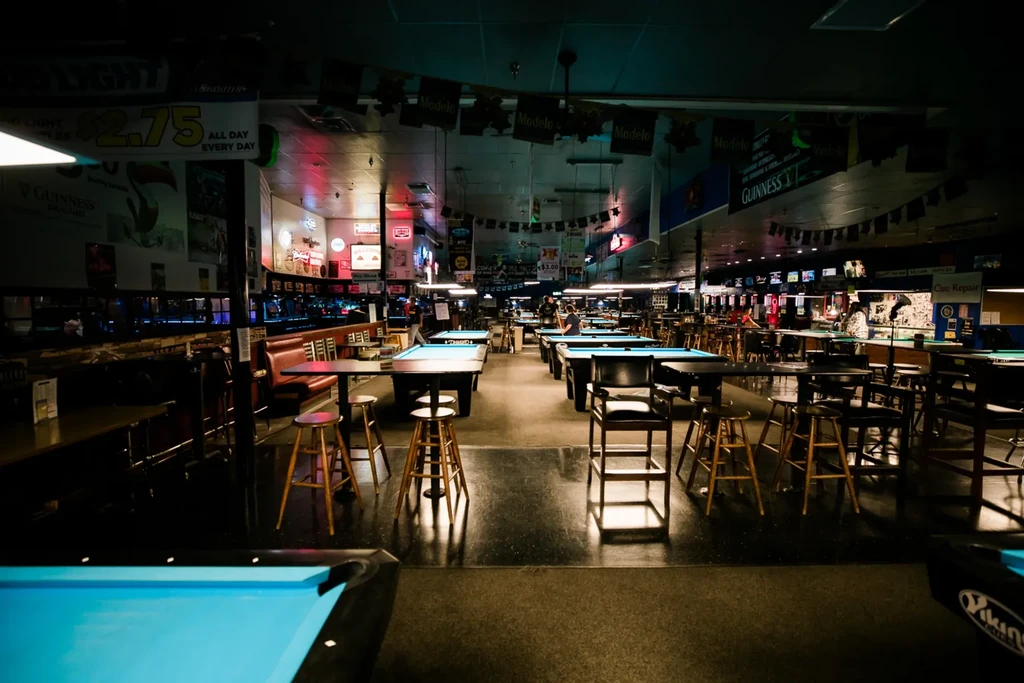 Spacious, dimly lit pool hall with many blue-felt tables and neon signs.