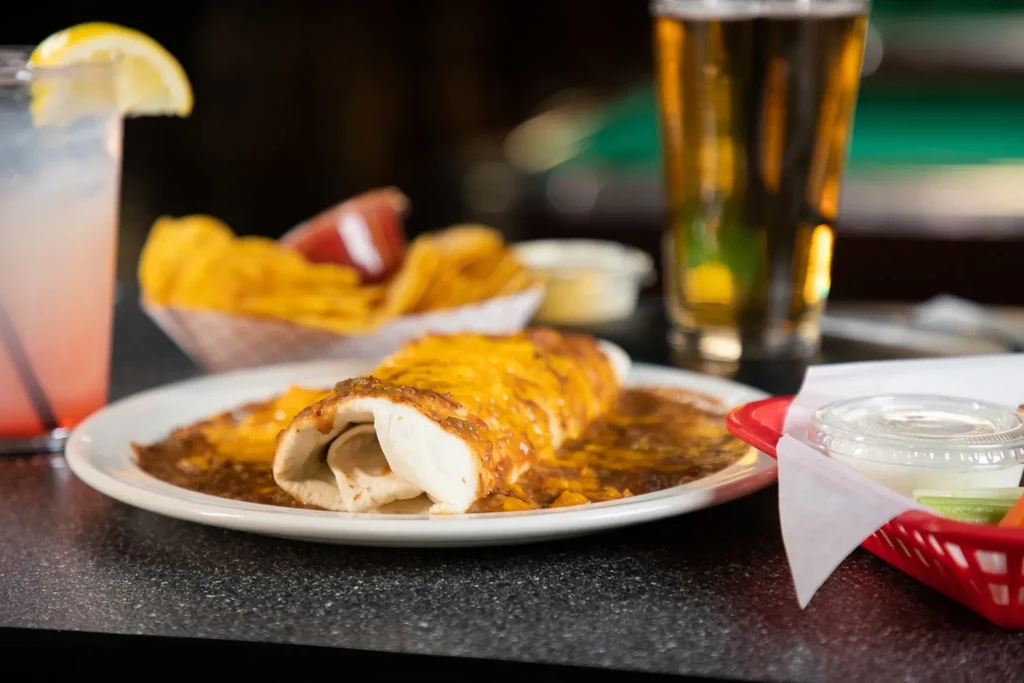 Burrito with cheese, drinks, chips, and veggies on a dark counter.