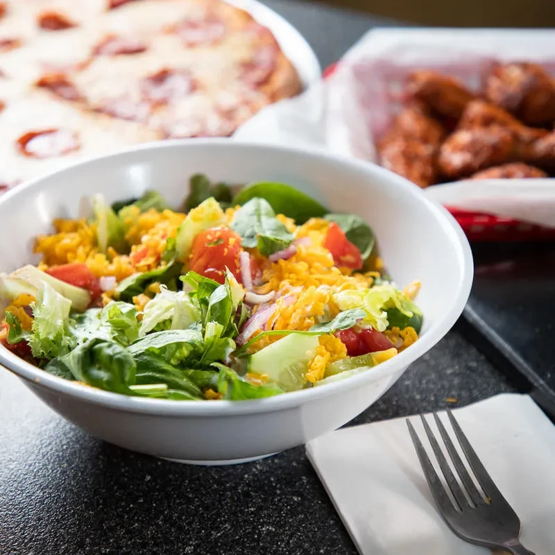 Fresh salad in white bowl, blurred pizza and chicken wings.
