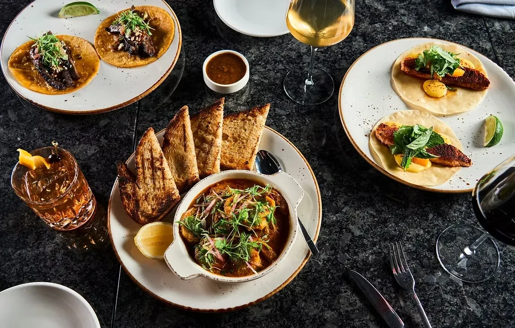 Overhead shot: dark table with gourmet tacos, stew, cocktails, and wine.