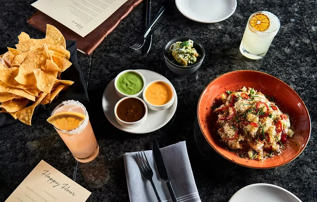 Overhead shot of restaurant table with chips, dips, drinks, and Happy Hour menu.