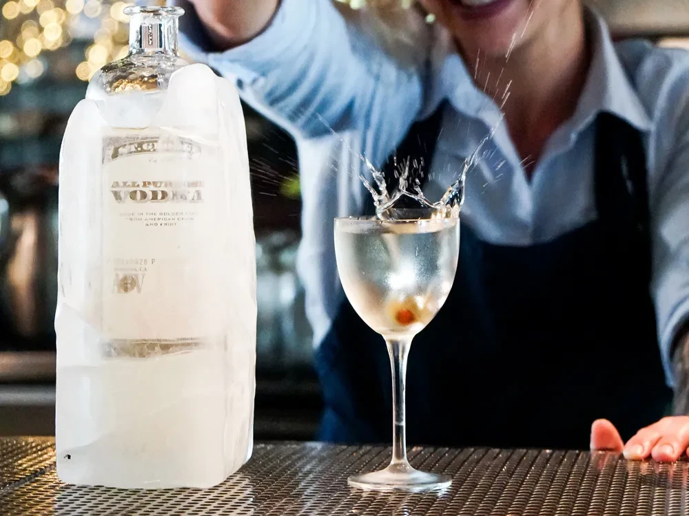 Ice-encrusted vodka bottle, splashing martini, and blurred bartender on a bar.