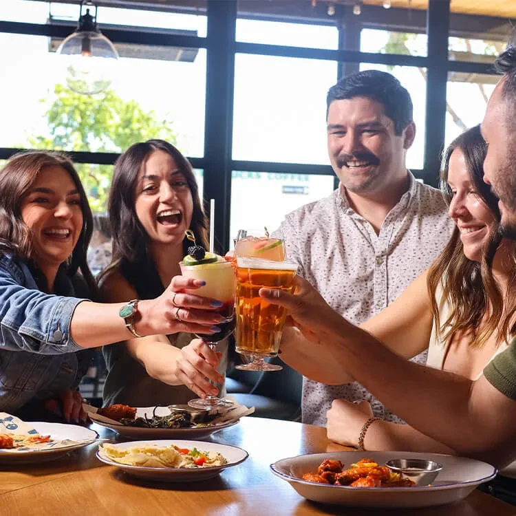 Five smiling people clinking drinks over a table of food.