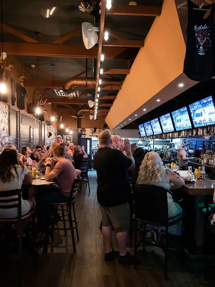 Lively restaurant and bar interior with patrons, sports TVs, warm lighting.