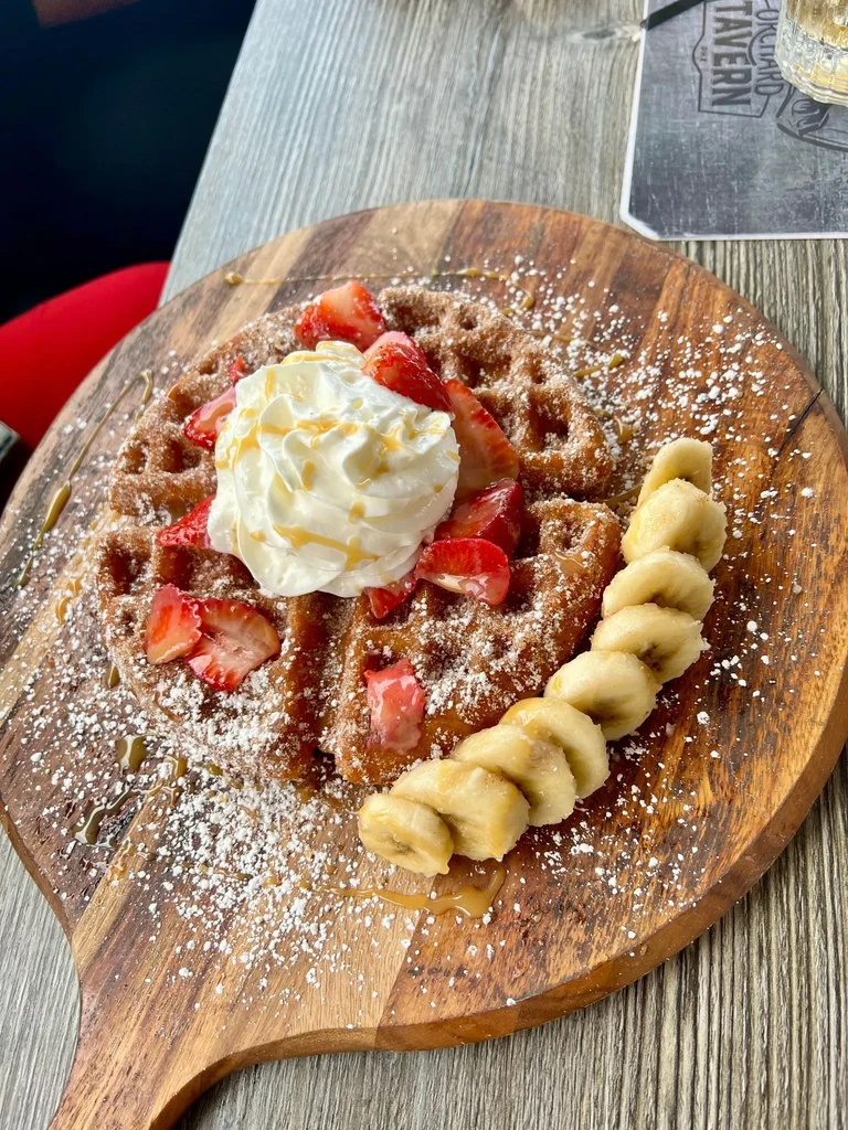 Waffle with whipped cream, strawberries, and bananas on a wooden board.