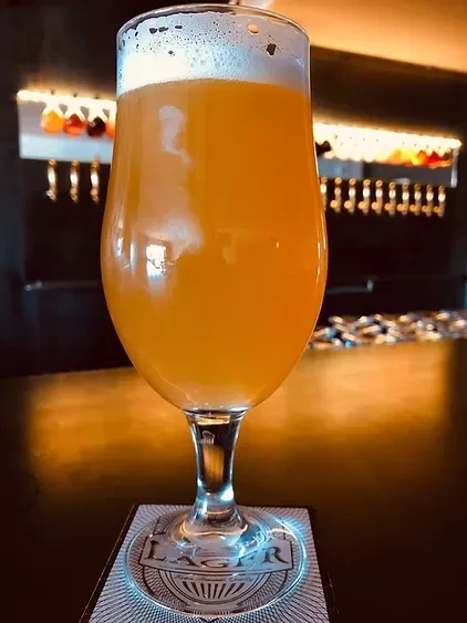Stemmed glass of cloudy orange-yellow beer with frothy head at a bar.