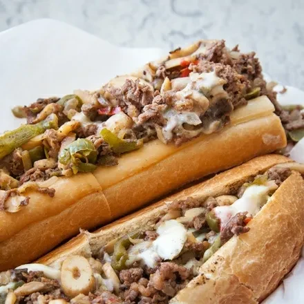 Close-up of two cheesesteak sandwiches with beef, melted cheese, and peppers.