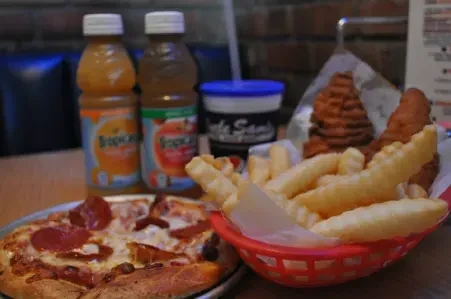 Pepperoni pizza, fries, chicken tenders, and juice bottles on a wooden table.