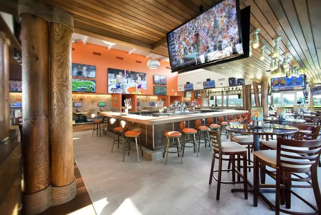 Spacious sports bar interior with central bar, orange walls, multiple TVs.