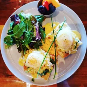 Two eggs Benedict, side salad, and fruit on a wooden table.