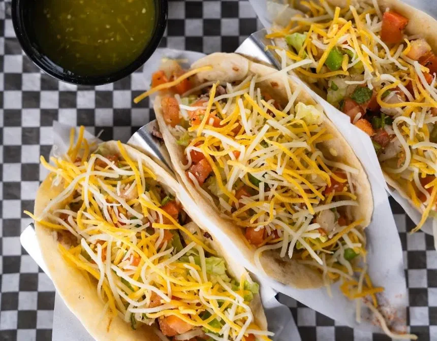 Overhead shot: three soft-shell tacos with cheese, lettuce, and green salsa.