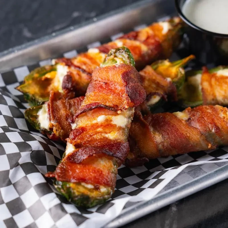 Bacon-wrapped jalapeño poppers with cheese and sauce on checkered paper.
