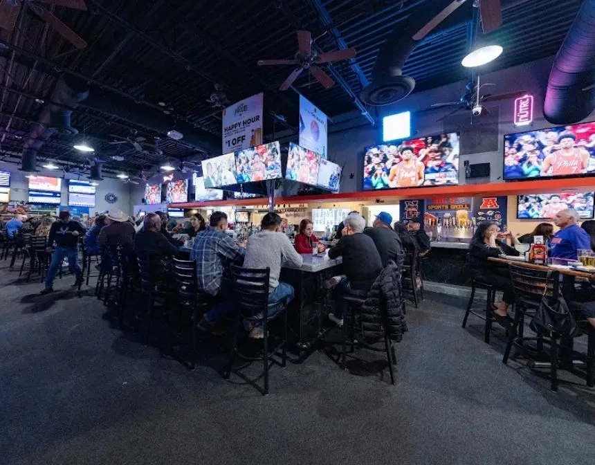 Lively sports bar interior with patrons, TVs showing sports, and neon signs.