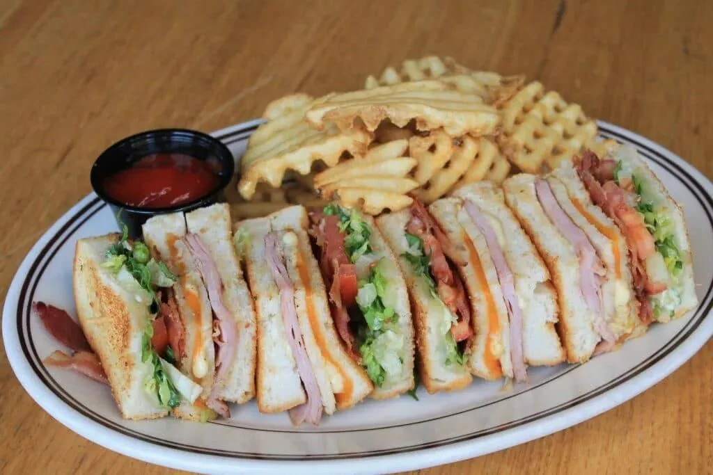 Triangular club sandwich, waffle fries, and ketchup on a wooden table.