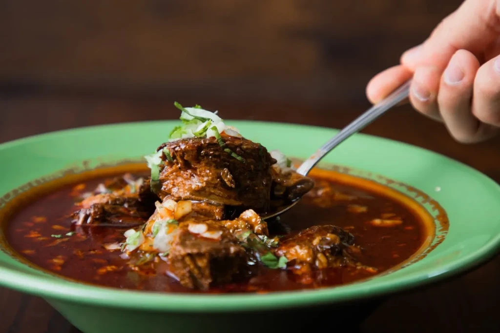 Reddish-brown meat stew with herbs in a green bowl, spoon lifting.
