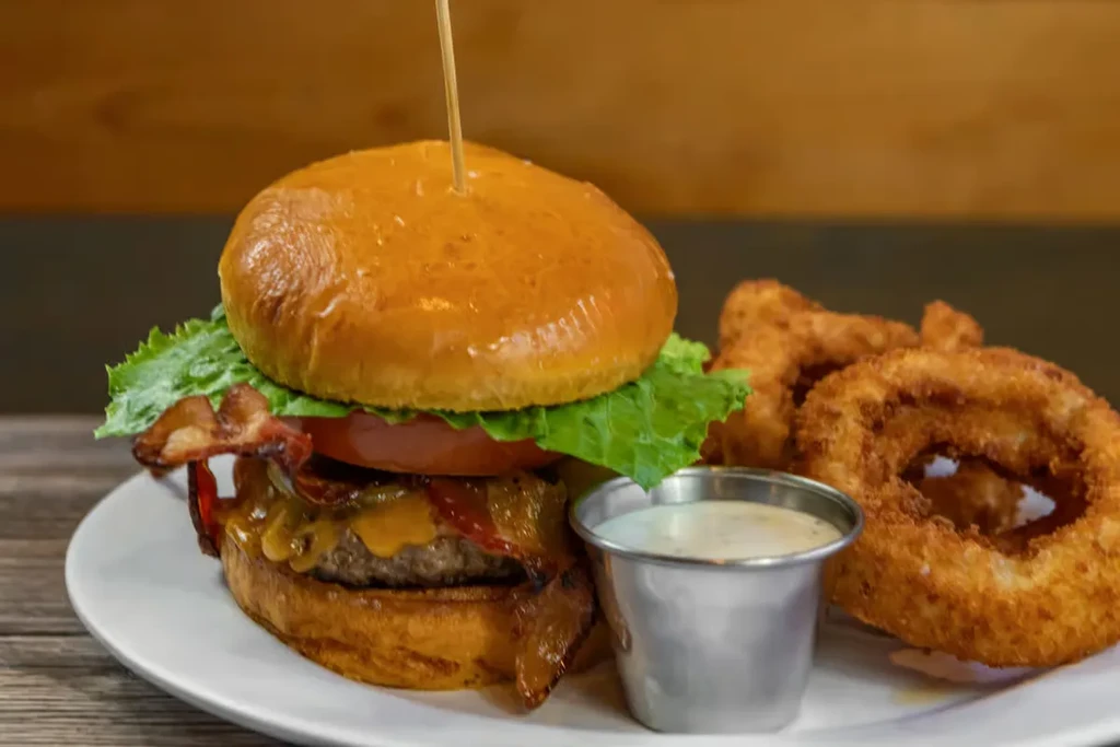 Gourmet burger with onion rings and dipping sauce on a white plate.