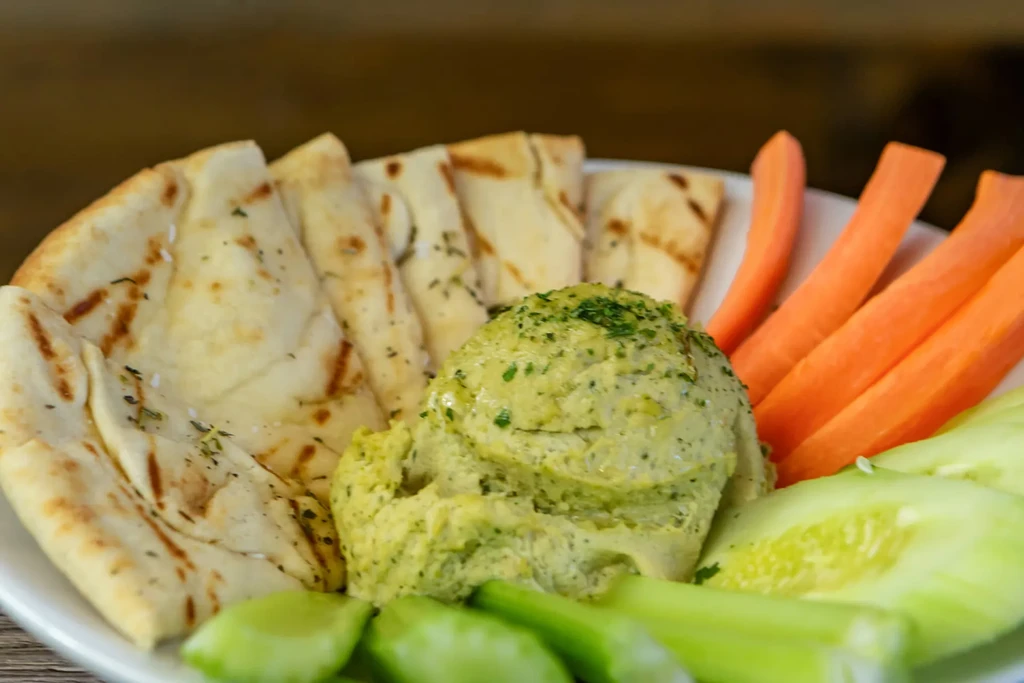 Light green hummus topped with herbs, pita bread, carrot, cucumber, and celery.