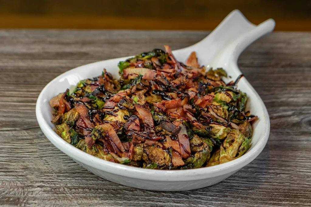 Charred roasted Brussels sprouts, crispy bacon, balsamic glaze in white bowl.